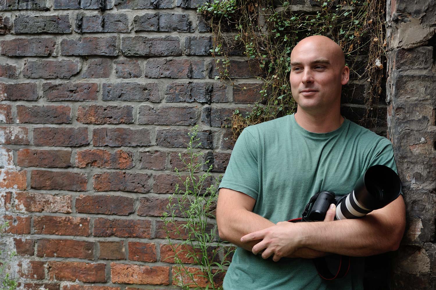 Jonathan of Studio Firin leaning against a brick wall holding his camera, Bristol